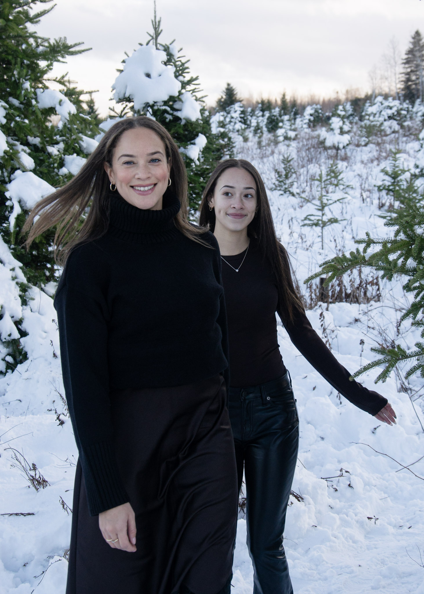 Two women walking in a snowy landscape, surrounded by evergreen trees, wearing black clothing and smiling.