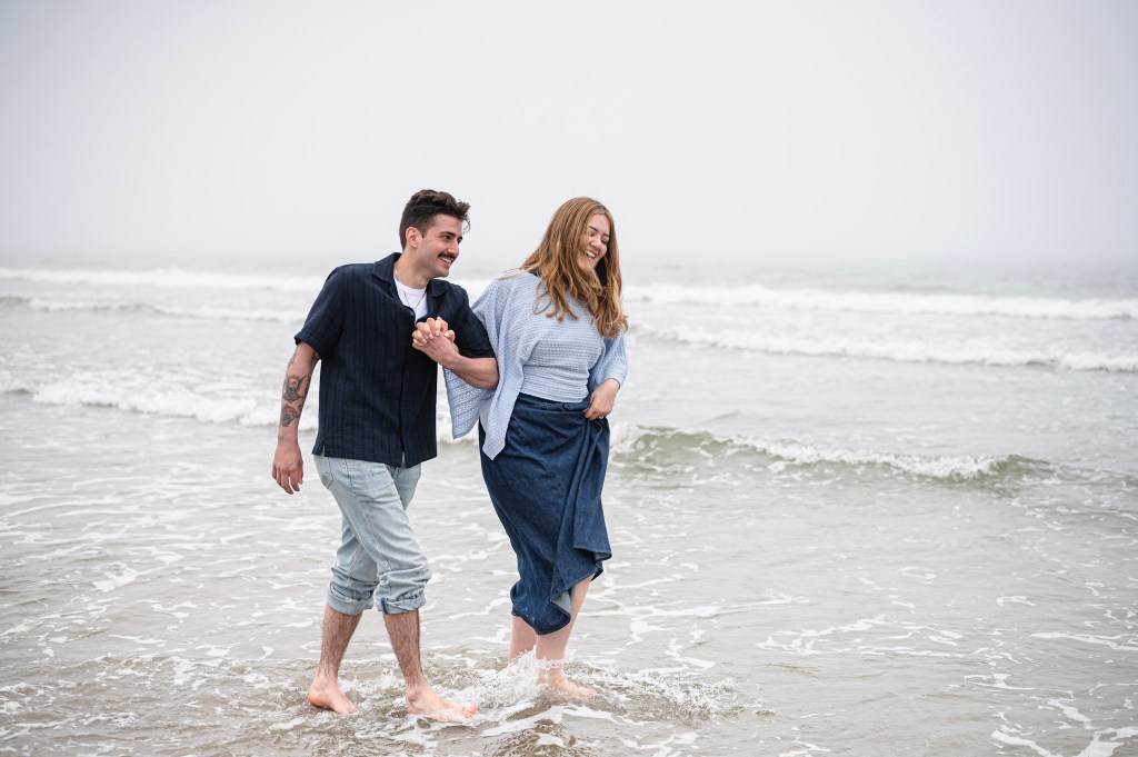 A couple joyfully walking hand in hand along the shore, with the ocean waves splashing around their feet on a foggy day.