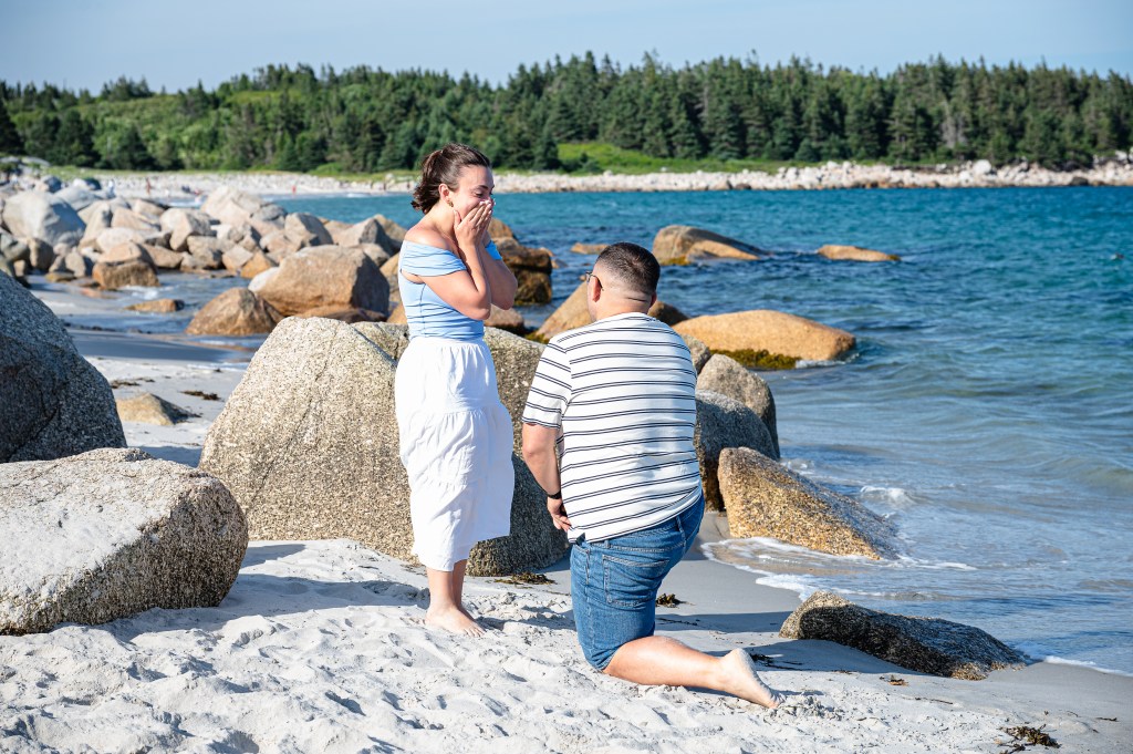 A man kneeling on the beach proposes to a surprised woman, who is covering her face with joy. They are surrounded by rocks and the ocean in the background.