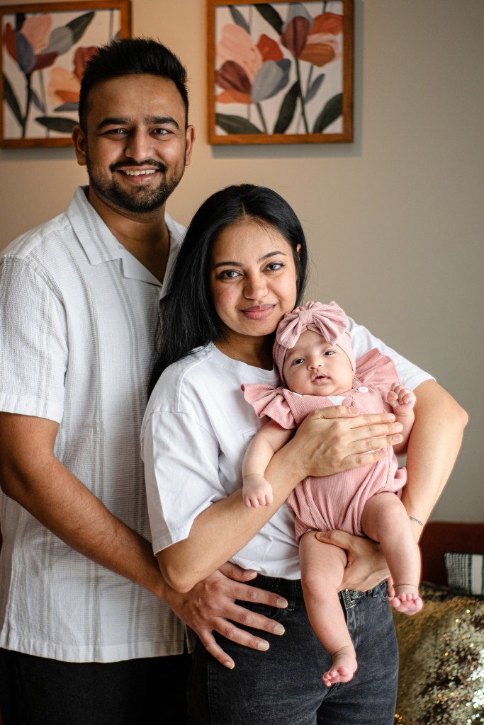 A family portrait featuring a smiling couple standing together, with the woman holding a baby. The woman is wearing a white shirt and the baby is dressed in a pink outfit with a matching headband. The man, dressed in a light-colored shirt, stands behind the woman with his hands on her waist. In the background, there are floral artworks on the walls.