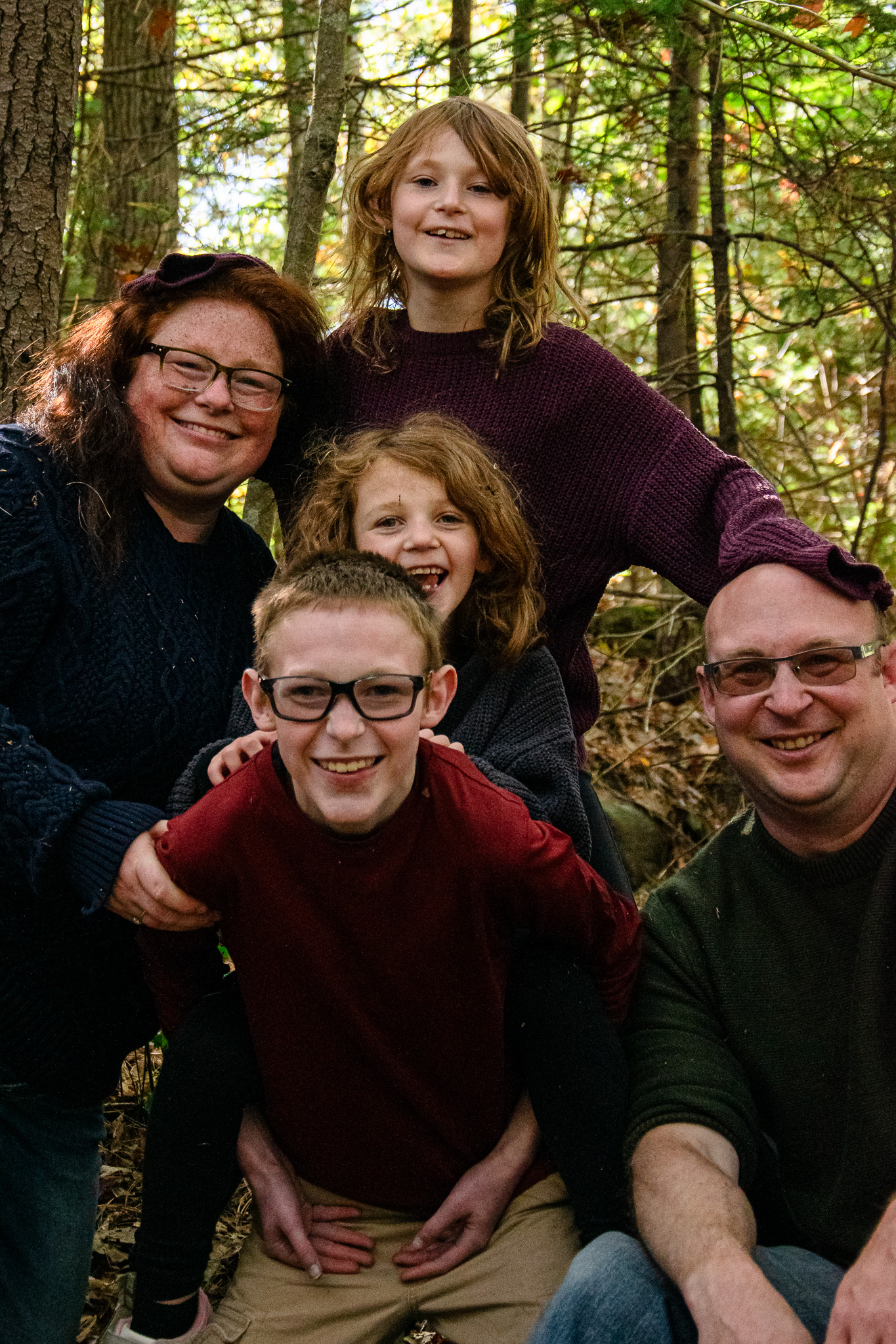 A family of five poses together in a wooded area, smiling and enjoying the moment. Two children are standing in the back while three others, including an adult couple, are seated in the front.