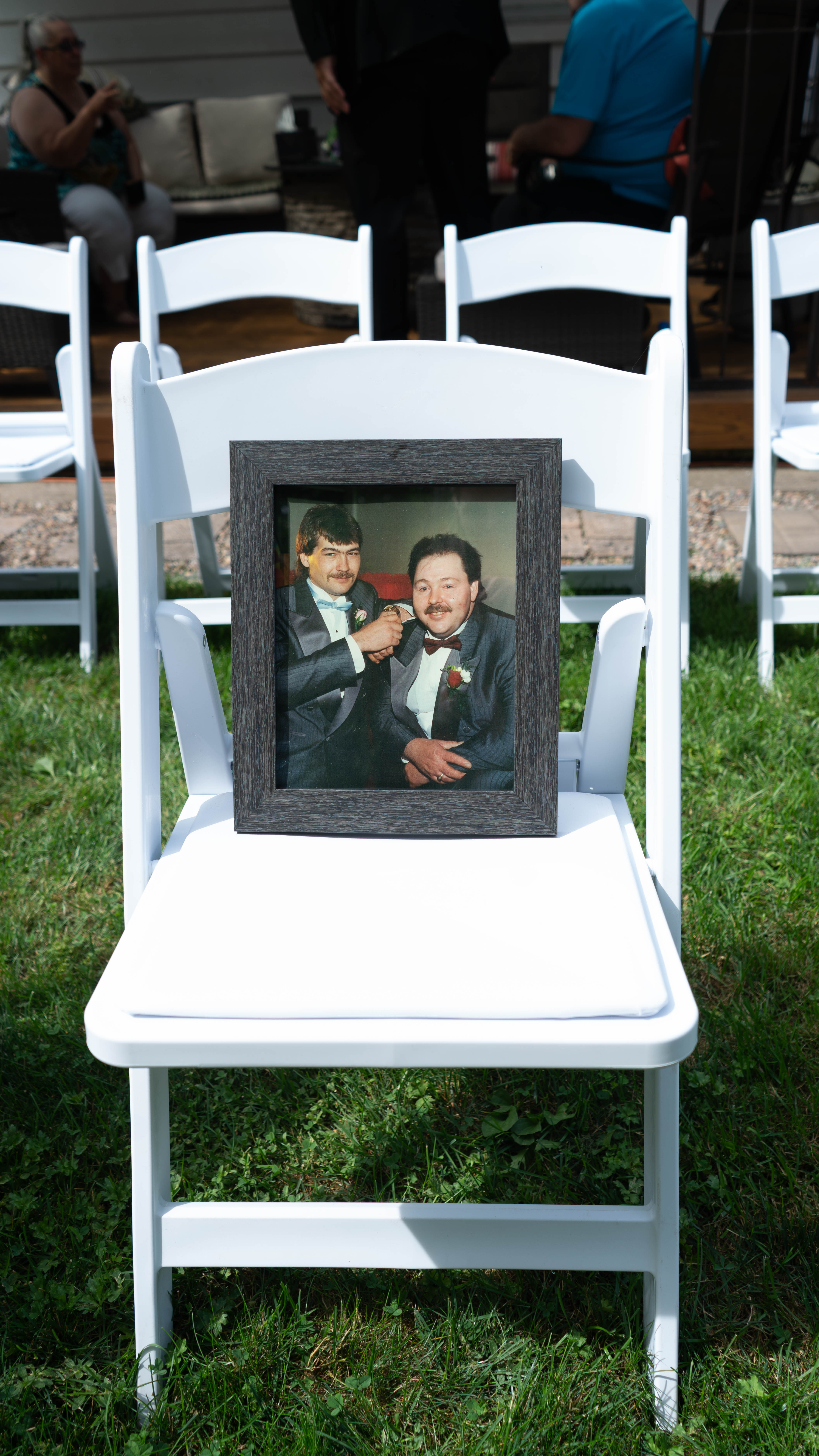 A framed photo of two men in formal attire sitting on a white chair placed on grass, with other empty chairs visible in the background.