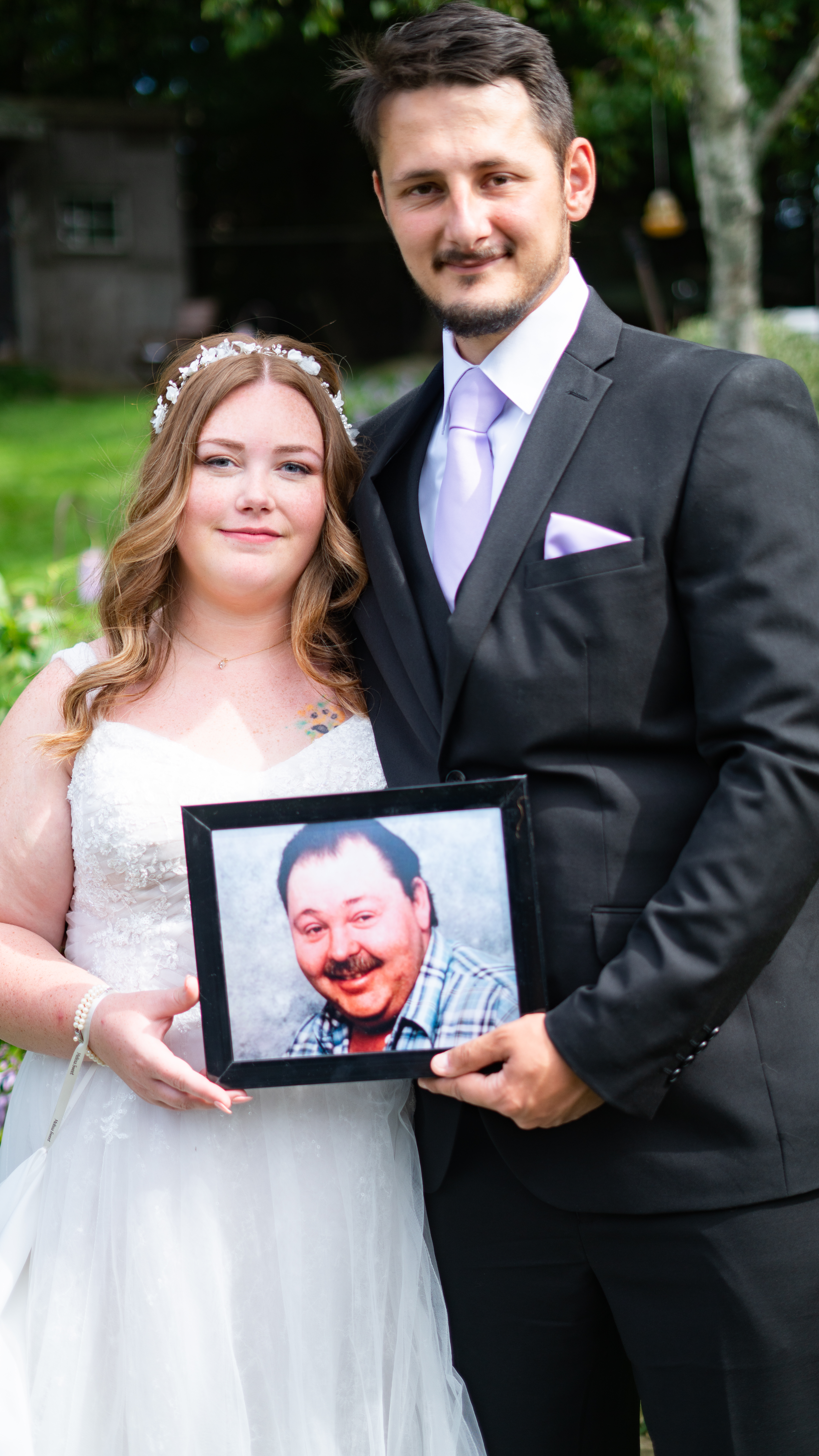 A bride and groom standing outdoors, holding a framed photograph of a man. The bride wears a white wedding dress and has a floral hairpiece, while the groom is dressed in a black suit with a light purple tie.