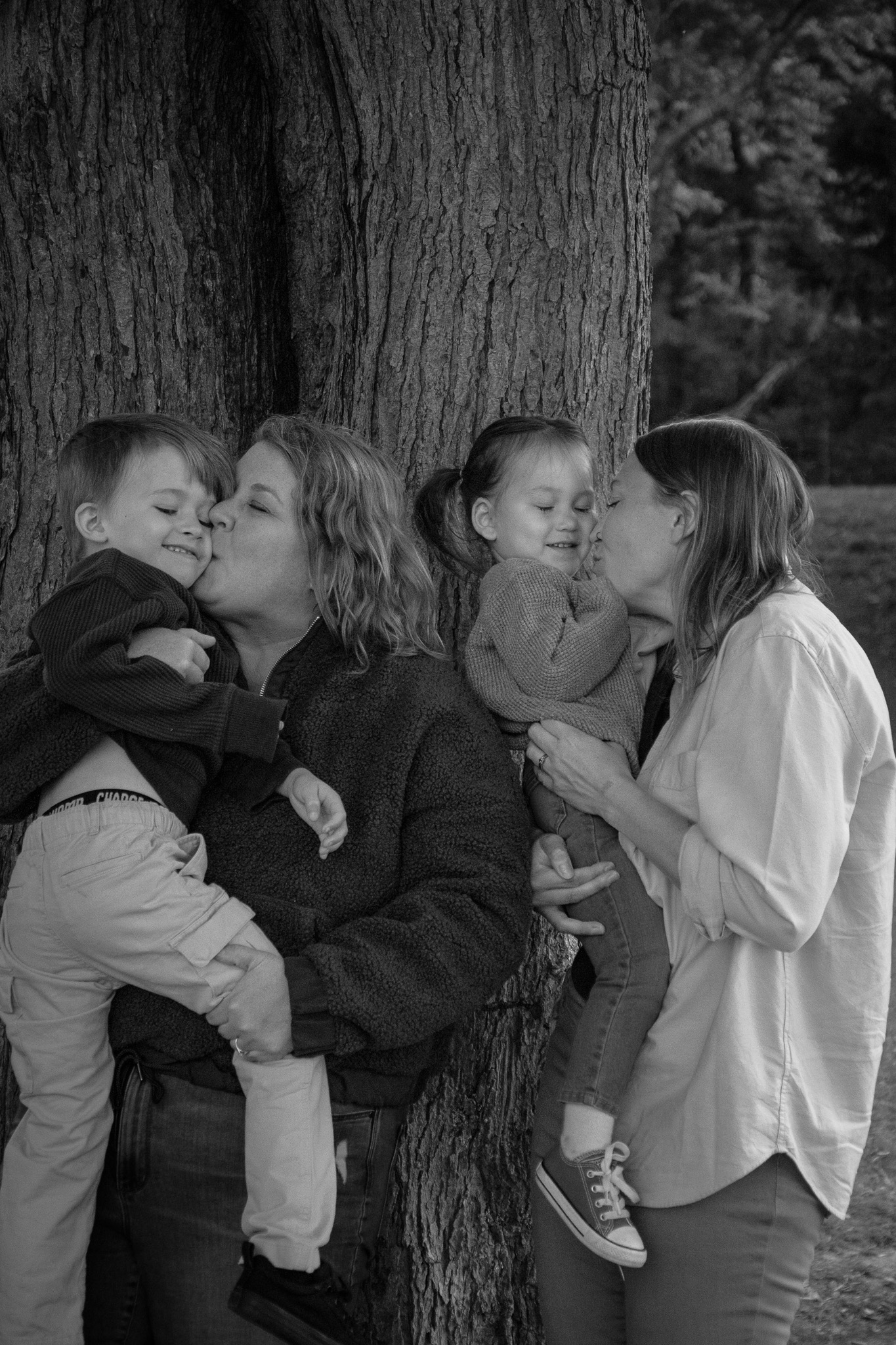 A black-and-white photo of two women kissing their children while standing against a tree. One woman holds a smiling boy, and the other woman holds a girl who is also smiling.