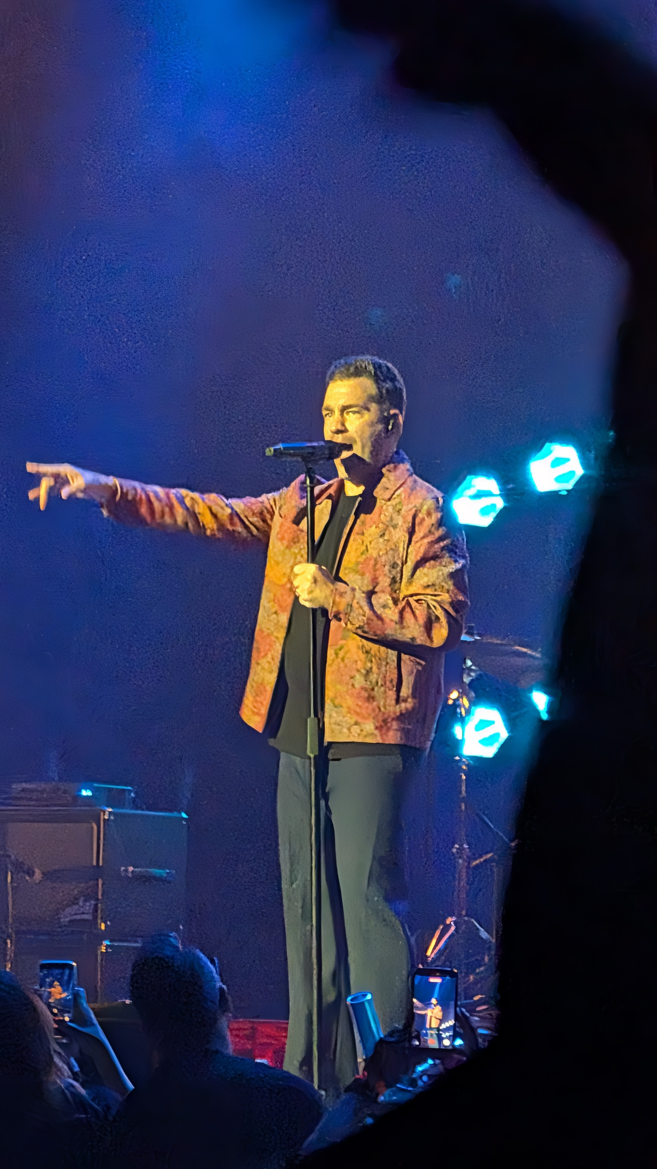 A singer performing on stage with a microphone, gesturing towards the audience, wearing a patterned jacket and standing in front of colorful stage lights.