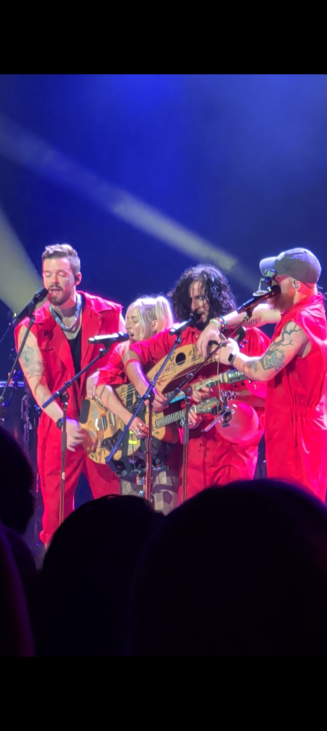 A group of four musicians, all wearing red outfits, perform together on stage. They are playing acoustic instruments and engaging with each other and the audience with enthusiasm.