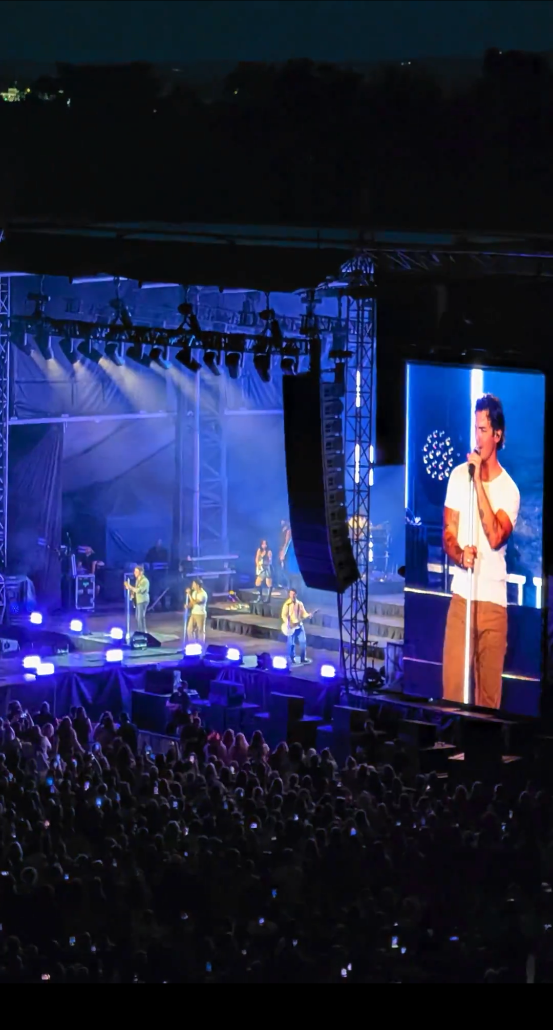 A live concert performance featuring a band on stage with colorful lighting, surrounded by an audience holding up phones. A large screen displays one of the band members singing.