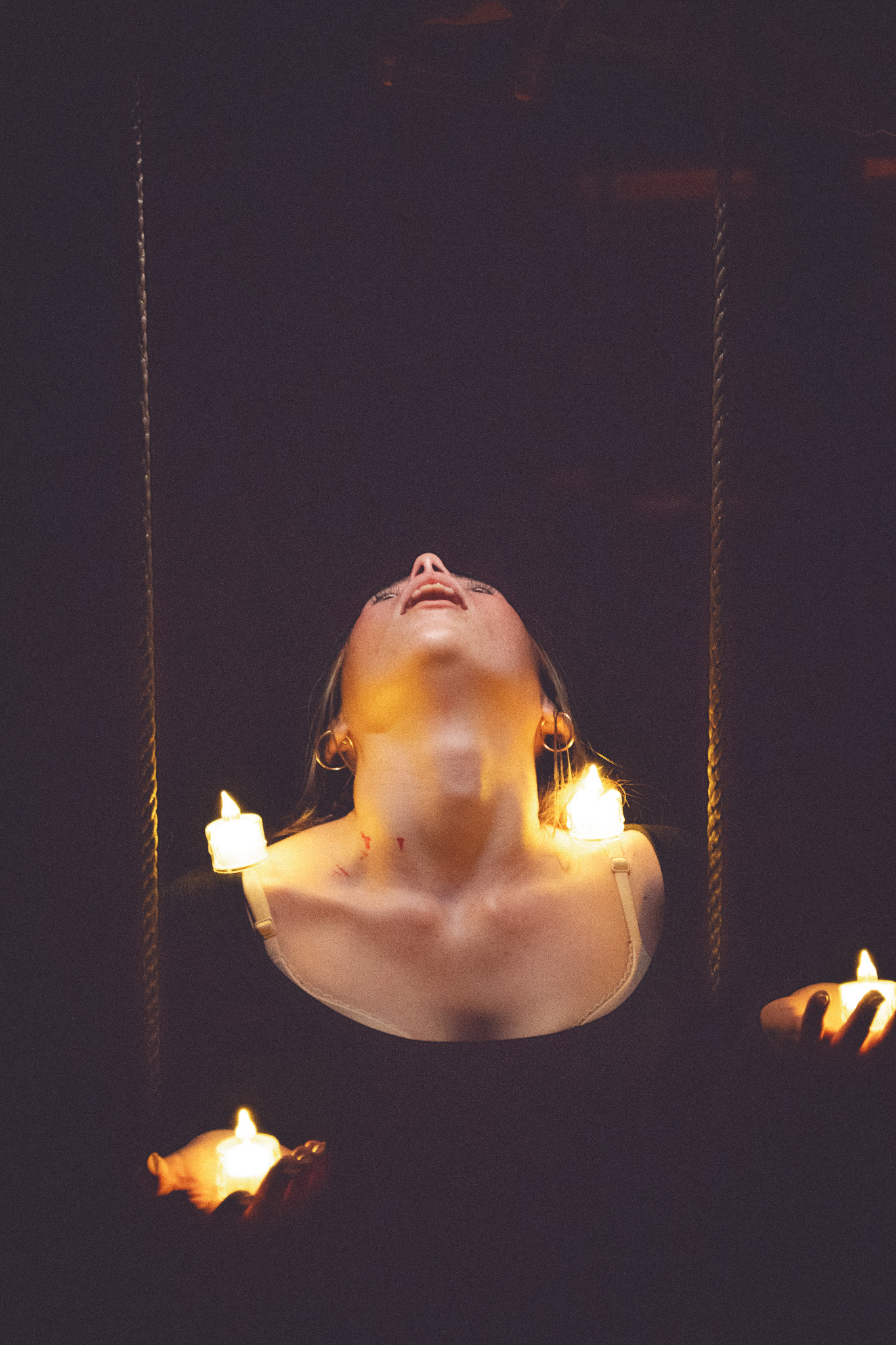 A person with their head tilted back, surrounded by glowing candles, with visible scratches on their neck, set against a dark background.