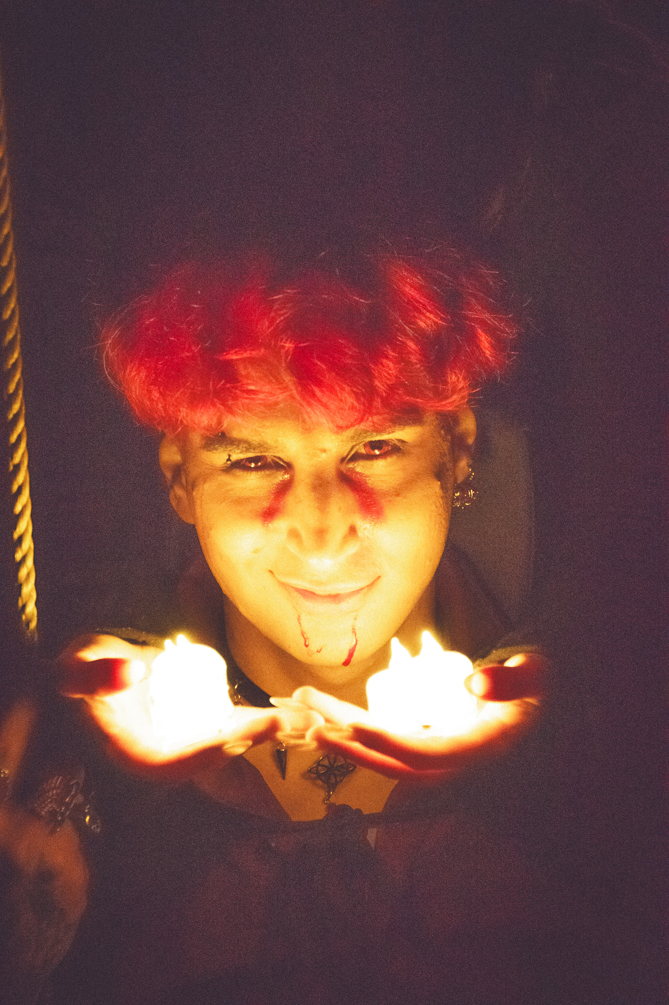 A person with red hair and dark clothing holds lit candles in their hands, with an intense expression and smeared makeup, creating a dramatic and mystical atmosphere.