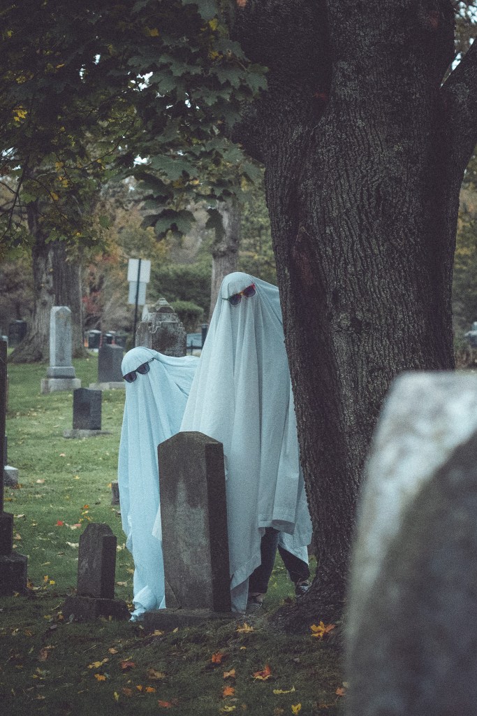 Two individuals dressed as ghosts in a graveyard, partially hidden behind a tree and gravestones, with a spooky autumn atmosphere.