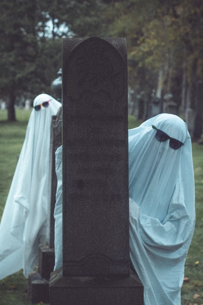 Two figures dressed as ghosts wearing sunglasses, hiding behind a gravestone in a cemetery.