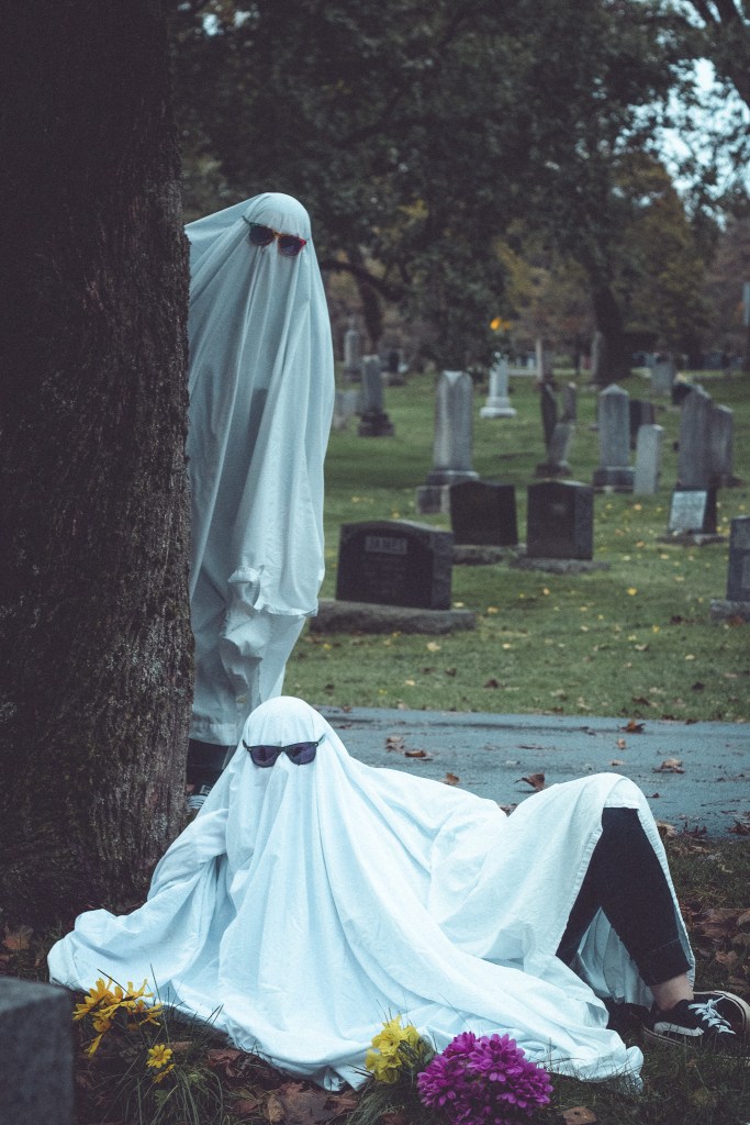 Two people dressed in white ghost costumes with sunglasses, one hiding behind a tree and the other sitting on the ground beside colorful flowers in a graveyard.