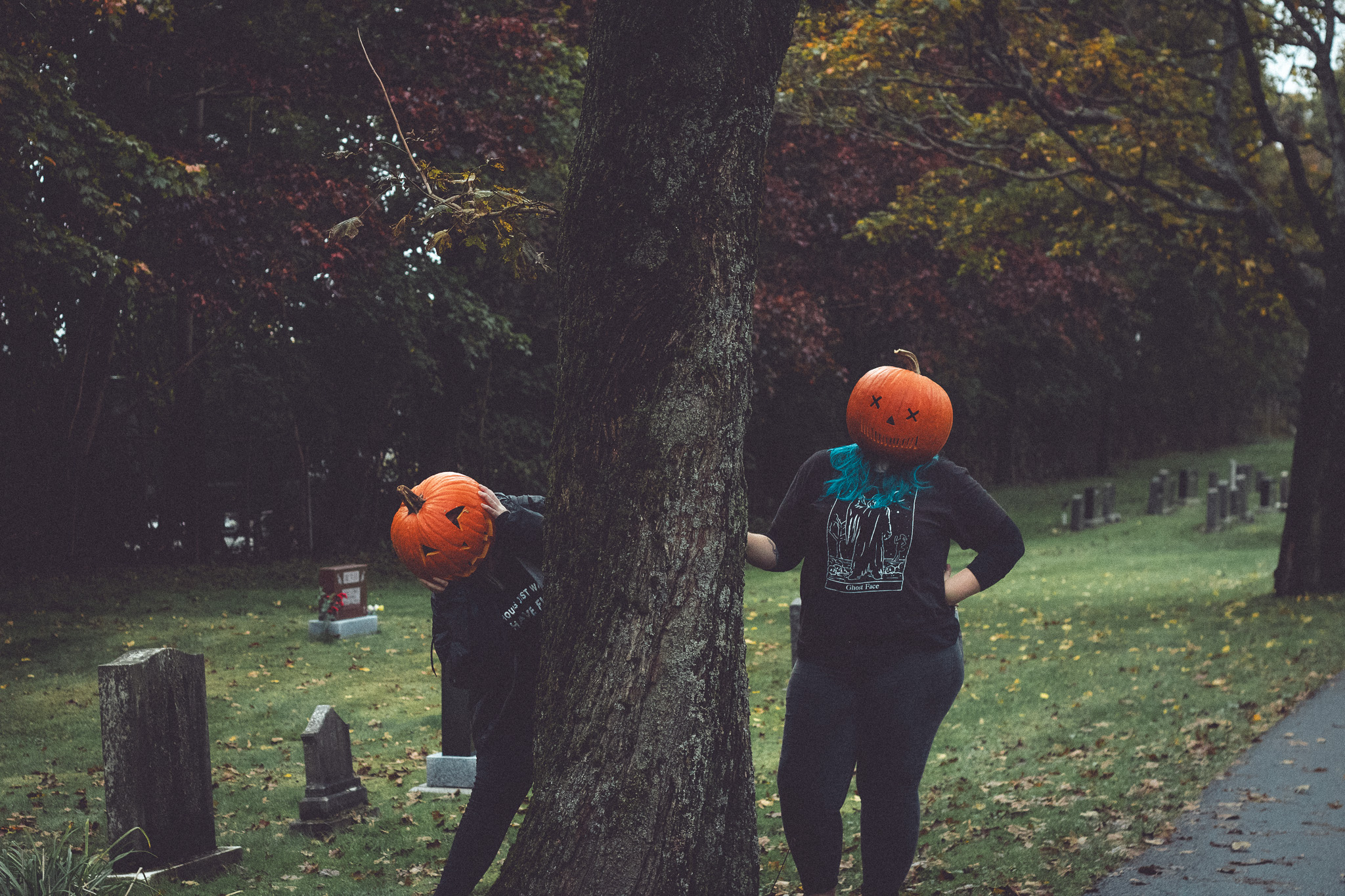 Two people with pumpkin heads in a cemetery, one peeking from behind a tree.