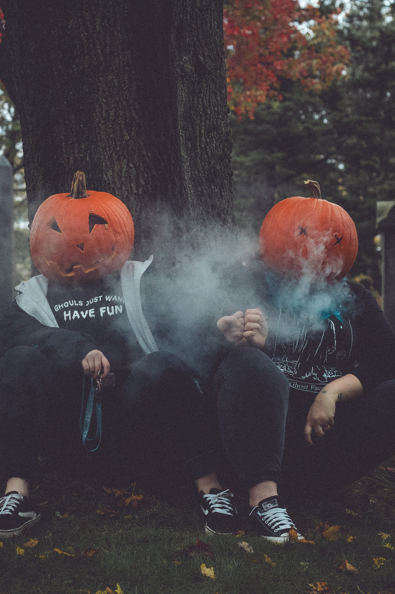 Two people sitting under a tree, wearing carved pumpkin heads, surrounded by autumn leaves and fog.